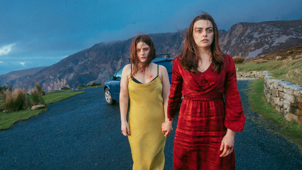 Two women with hints of blood on their clothes and faces hold hands in the centre of the image. They are walking away from a car parked on a rural road with rocky hills in the background.